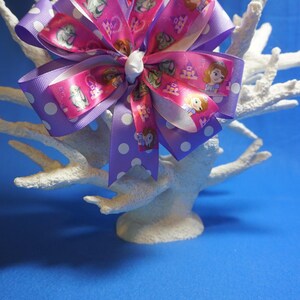 May include: A pink and purple bow with white polka dots and a princess character print. The bow is tied with a white ribbon and is displayed on a white coral.