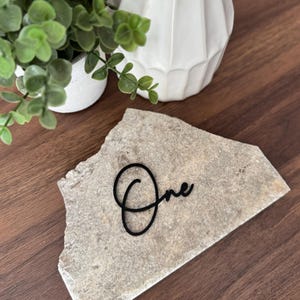K&ouml;nnte beinhalten: Eine Tischkarte aus Naturstein mit dem Wort "One" in schwarzer Schreibschrift. Der Stein ist hellgrau und liegt auf einer dunkelbraunen Holzoberfl&auml;che. Eine kleine wei&szlig;e Vase und eine Topfpflanze befinden sich im Hintergrund.