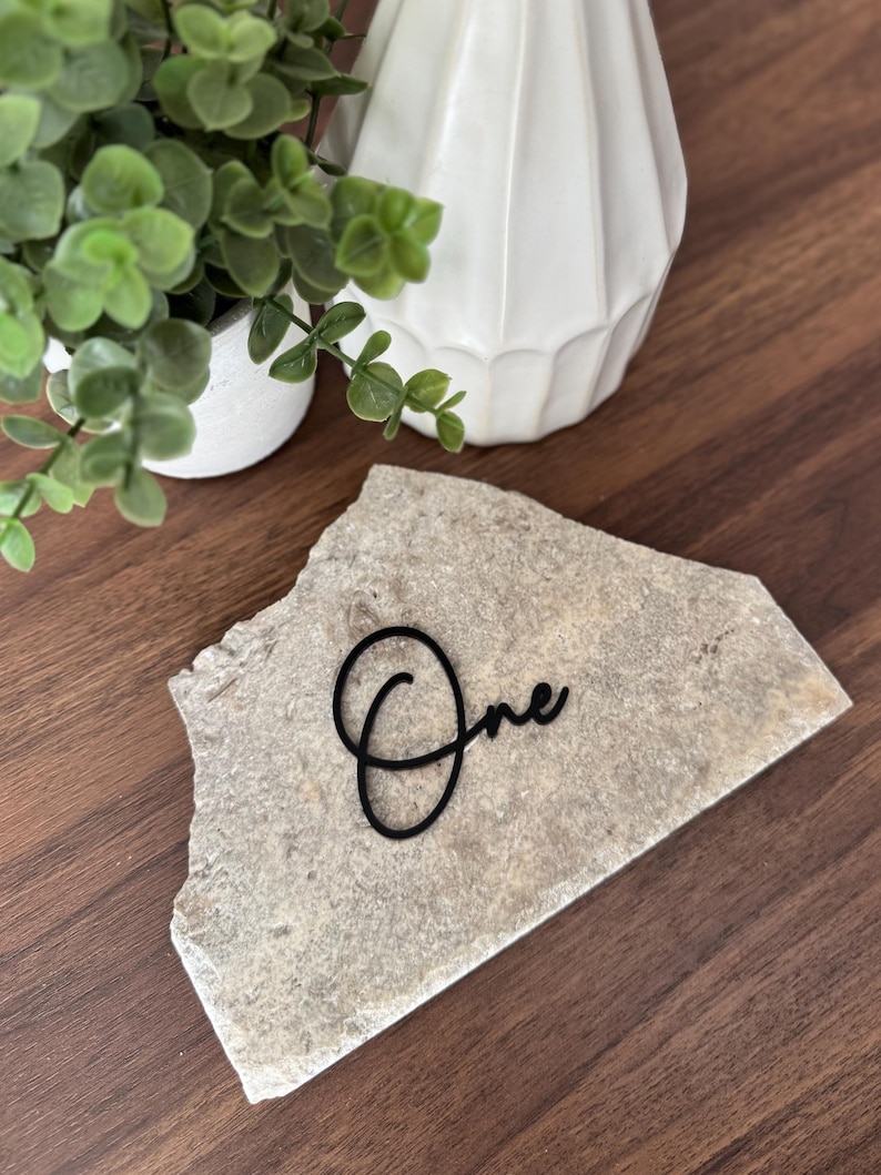 K&ouml;nnte beinhalten: Eine Tischkarte aus Naturstein mit dem Wort "One" in schwarzer Schreibschrift. Der Stein ist hellgrau und liegt auf einer dunkelbraunen Holzoberfl&auml;che. Eine kleine wei&szlig;e Vase und eine Topfpflanze befinden sich im Hintergrund.