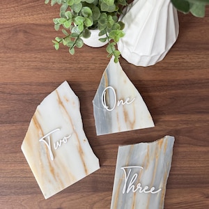 K&ouml;nnte beinhalten: Drei Tischkarten mit Marmor-Tischnummern und wei&szlig;er Schrift. Die Karten sind in Grau-, Wei&szlig;- und Goldt&ouml;nen gehalten. Die Zahlen sind "Eins", "Zwei" und "Drei". Eine wei&szlig;e Vase und eine kleine Topfpflanze befinden sich im Hintergrund.