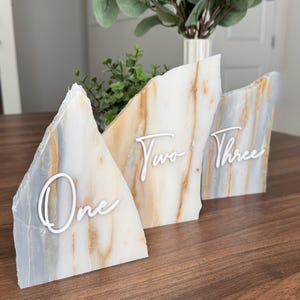 K&ouml;nnte beinhalten: Drei dekorative Tischnummernschilder. Jedes Schild ist ein einzigartiges, unregelm&auml;&szlig;ig geformtes St&uuml;ck Stein mit wei&szlig;en Schriftnummern: "One", "Two" und "Three". Der Stein hat ein marmoriertes Aussehen mit Wei&szlig;-, Grau- und Goldt&ouml;nen. Die Schilder stehen auf einer dunklen Holzoberfl&auml;che.