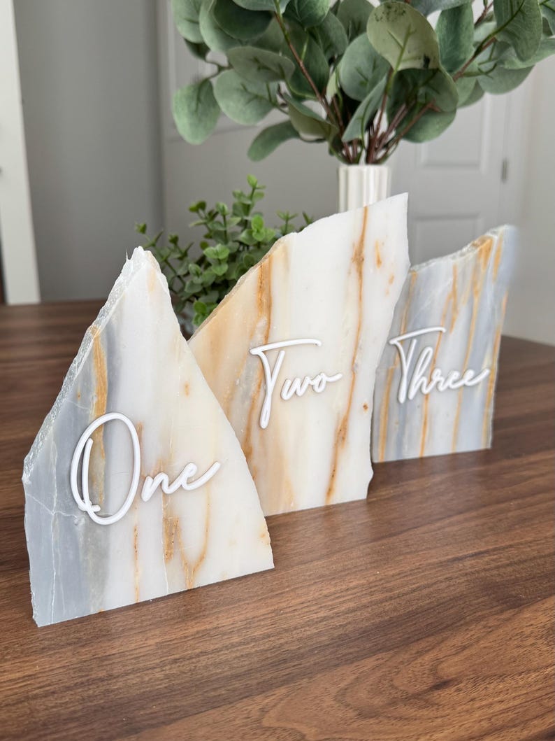K&ouml;nnte beinhalten: Drei dekorative Tischnummernschilder. Jedes Schild ist ein einzigartiges, unregelm&auml;&szlig;ig geformtes St&uuml;ck Stein mit wei&szlig;en Schriftnummern: "One", "Two" und "Three". Der Stein hat ein marmoriertes Aussehen mit Wei&szlig;-, Grau- und Goldt&ouml;nen. Die Schilder stehen auf einer dunklen Holzoberfl&auml;che.