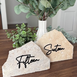 K&ouml;nnte beinhalten: Zwei Tischkartenhalter aus Naturstein, einer mit dem Wort "Fifteen" und der andere mit "Sixteen" in schwarzer Schrift. Die Steine sind beige und stehen auf einer dunklen Holzoberfl&auml;che, mit Gr&uuml;n und einer wei&szlig;en Vase im Hintergrund.