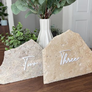 K&ouml;nnte beinhalten: Zwei Tischkarten aus Stein mit wei&szlig;em Schreibschrifttext. Der Stein hat eine hellbeige Farbe. Das Wort "Two" steht auf dem linken Stein und das Wort "Three" auf dem rechten Stein. Eine wei&szlig;e Vase mit Gr&uuml;nzeug befindet sich im Hintergrund.