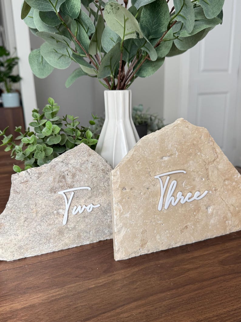 K&ouml;nnte beinhalten: Zwei Tischkarten aus Stein mit wei&szlig;em Schreibschrifttext. Der Stein hat eine hellbeige Farbe. Das Wort "Two" steht auf dem linken Stein und das Wort "Three" auf dem rechten Stein. Eine wei&szlig;e Vase mit Gr&uuml;nzeug befindet sich im Hintergrund.