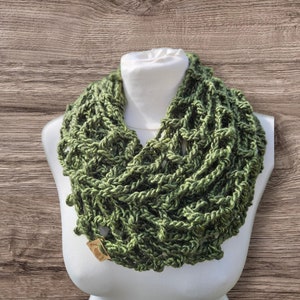 May include: A green crocheted infinity scarf on a white mannequin. The scarf features an open, textured weave and a tag. The background is a wooden surface.