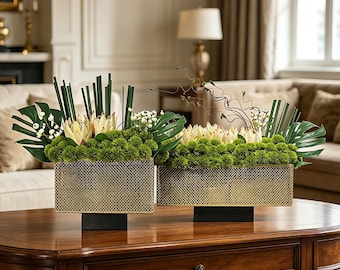 May include: Two rectangular gold-toned metal mesh containers with black bases, each filled with a floral arrangement featuring green moss-like filler, white protea-style flowers, large green monstera leaves, and tall green stems.