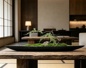 May include: A long, narrow, black, boat-shaped decorative tray featuring a textured, sunburst-like carved pattern. The tray contains a bed of vibrant green moss, a single grey stone, and a delicate, thin, curved branch with small green leaves.