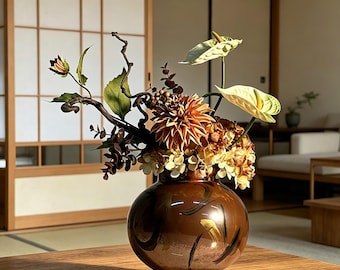 May include: A round, glossy brown ceramic vase with dark brushstroke patterns, containing an arrangement of cream-colored anthurium flowers, a large orange dahlia, dried hydrangea clusters, and dark, twisted branches with green leaves.