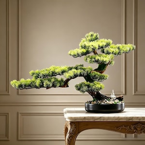 Puede incluir: Un bonsái artificial con follaje verde vibrante en forma de agujas y un tronco texturizado de color marrón oscuro. El árbol está plantado en una maceta redonda de cerámica negra con piedras claras y una pequeña figura blanca de Buda.