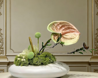 May include: A white, oval-shaped ceramic vase holds an arrangement of flowers and greenery. The arrangement includes a large, pink and green anthurium, green spherical flowers, and various green foliage. The vase sits on a marble-like surface.