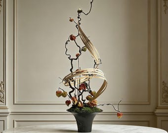 May include: A floral arrangement in a black flared vase featuring dark, twisted branches, orange physalis husks, and curved, light-colored wooden strips. The base of the arrangement is covered in green moss, creating a vertical, sculptural display.