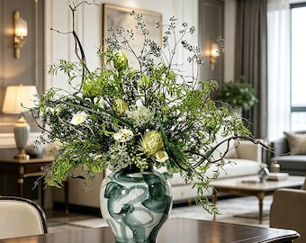 May include: A ceramic vase with a white base and dark green abstract brushstroke patterns, filled with a large arrangement of green foliage, thin dark branches, and light green protea-like flowers, resting on a dark wooden table surface.