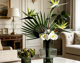 May include: A tall, dark green glass vase containing bird of paradise flowers, white orchids, and green palm leaves, positioned next to a shorter, matching glass bowl filled with green artichokes on a white marble surface.