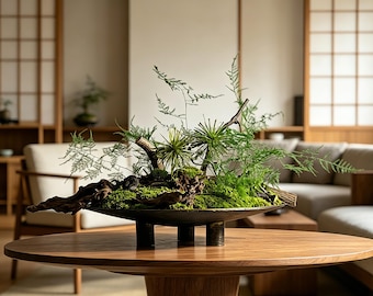 May include: A shallow, dark metal bowl with three short cylindrical legs, containing a lush arrangement of vibrant green moss, delicate fern fronds, and textured pieces of dark driftwood, creating a natural, organic centerpiece display.