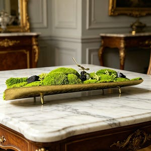 Könnte beinhalten: Ein dekoratives Centerpiece mit einer langen, goldfarbenen Metallschale, gefüllt mit leuchtend grünem Moos, kleinen schwarzen Steinen und einer goldfarbenen Insektenfigur. Die Schale wird von kurzen, goldfarbenen Beinen getragen. Die Anordnung steht auf einem Marmortisch.