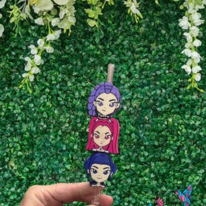 May include: A clear badge reel featuring three cartoon girl faces with purple, pink, and blue hair. The faces have black eyes and are smiling. The badge reel is held in a hand against a green background with white flowers.