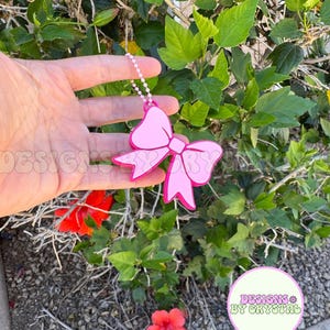 May include: A pink bow-shaped keychain with a white chain. The keychain is held in front of green foliage with red flowers. The text "DESIGNS BY CRYSTAL" is visible in a circular logo.