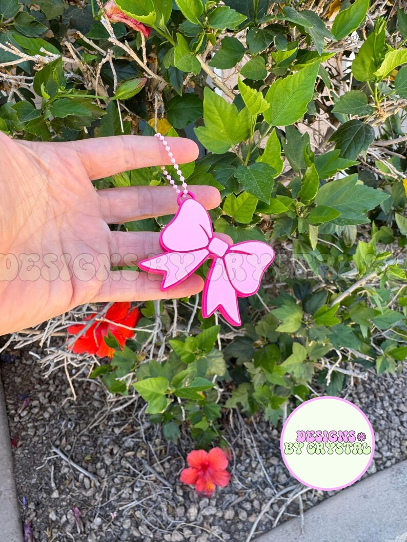 May include: A pink bow-shaped keychain with a white chain. The keychain is held in front of green foliage with red flowers. The text "DESIGNS BY CRYSTAL" is visible in a circular logo.