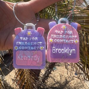 May include: Two backpack-shaped luggage tags, one purple and one pink, with the text "TAP FOR EMERGENCY CONTACT". The purple tag says "Kennedy" and the pink tag says "Brooklyn". Both tags have daisy accents and a silver loop.