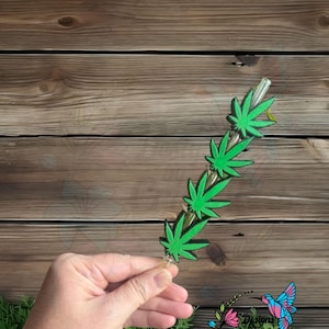 May include: A clear glass smoking pipe with a series of green cannabis leaf designs. The pipe is held against a wooden background. The pipe is approximately 6 inches long.