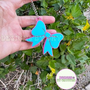 May include: A turquoise bow-shaped charm with a pink outline, hanging from a silver chain. The charm is held against a backdrop of green foliage. The text "DESIGNS BY CRYSTAL" is visible in a circular logo.