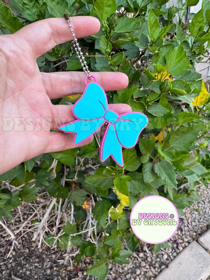 May include: A turquoise bow-shaped charm with a pink outline, hanging from a silver chain. The charm is held against a backdrop of green foliage. The text "DESIGNS BY CRYSTAL" is visible in a circular logo.
