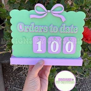May include: A light green rectangular sign with scalloped edges and a lavender bow at the top. The sign features the text "Orders to date" in lavender lettering above three lavender squares, each displaying a white number, currently showing 100.
