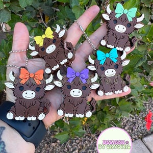 May include: A collection of six brown Highland cow keychains with white horns and feet, each adorned with a different colored bow: yellow, orange, purple, blue, and green. The keychains are held by a hand against a backdrop of green foliage.