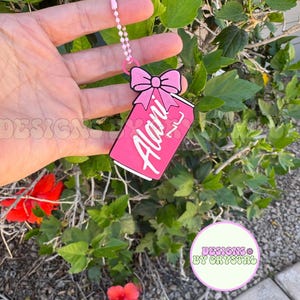 May include: A pink tag-shaped keychain with a bow at the top and the name "Alani" in white script. The keychain is attached to a pink ball chain. The background features green foliage and a few red flowers.