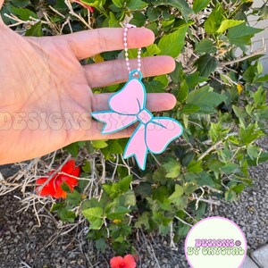 May include: A pink bow-shaped keychain with a light blue outline and a silver chain. The bow has a decorative design on the front. The text "DESIGNS BY CRYSTAL" is visible in a circular logo.