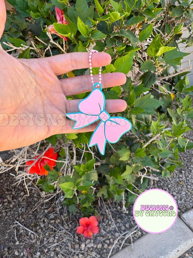 May include: A pink bow-shaped keychain with a light blue outline and a silver chain. The bow has a decorative design on the front. The text "DESIGNS BY CRYSTAL" is visible in a circular logo.