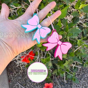 May include: Two pink bow-shaped keychains with white beaded chains. One bow has a teal outline, the other is solid pink. The keychains are held in front of a green leafy background with red flowers. The text "DESIGNS BY CRYSTAL" is in a circle.