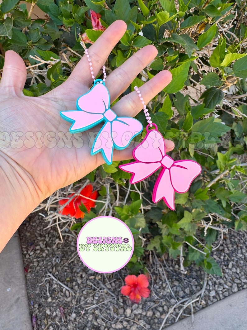 May include: Two pink bow-shaped keychains with white beaded chains. One bow has a teal outline, the other is solid pink. The keychains are held in front of a green leafy background with red flowers. The text "DESIGNS BY CRYSTAL" is in a circle.