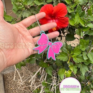 May include: A pink bow-shaped keychain with a light blue outline hangs from a silver chain. The keychain is held in front of a vibrant red hibiscus flower and lush green foliage. The text "DESIGNS BY CRYSTAL" is visible.