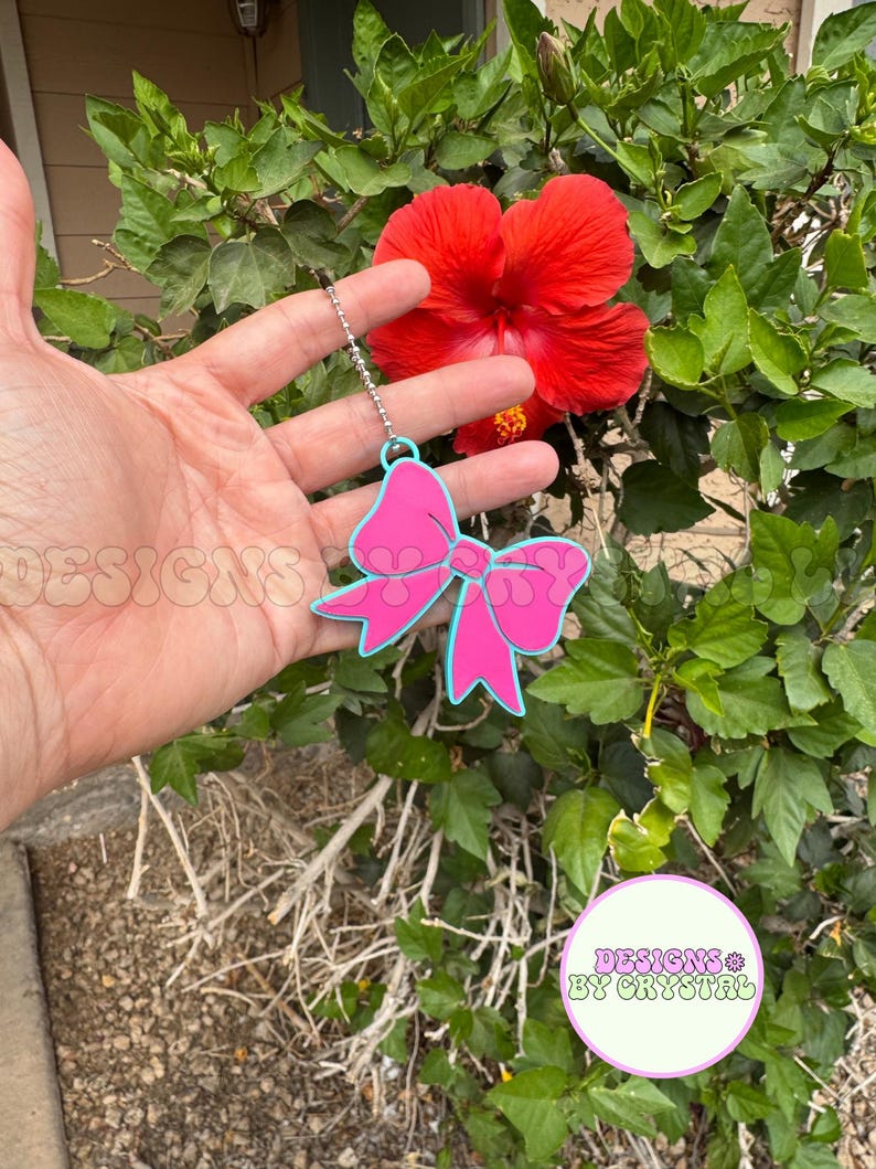 May include: A pink bow-shaped keychain with a light blue outline hangs from a silver chain. The keychain is held in front of a vibrant red hibiscus flower and lush green foliage. The text "DESIGNS BY CRYSTAL" is visible.
