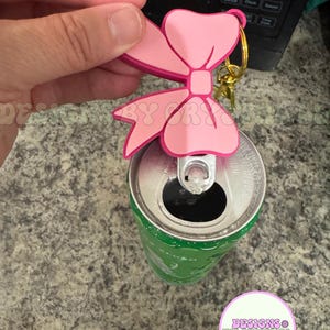May include: A pink bow-shaped keychain with a gold-colored ring is attached to a green soda can. The bow has a darker pink outline. The can is open. The text "DESIGNS BY CRYSTAL" is in a circle.