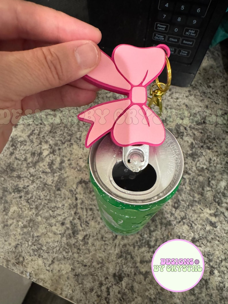 May include: A pink bow-shaped keychain with a gold-colored ring is attached to a green soda can. The bow has a darker pink outline. The can is open. The text "DESIGNS BY CRYSTAL" is in a circle.
