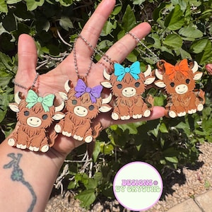 May include: Four Highland cow keychains with different colored bows: green, purple, blue, and orange. Each keychain is brown with a smiling face and a silver chain. The text "DESIGNS BY CRYSTAL" is in a circle in the lower right corner.
