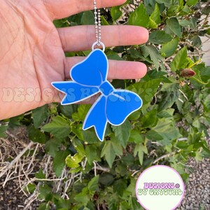 May include: A blue bow-shaped charm with a white outline, hanging from a silver ball chain. The charm is held in front of a green leafy background. The text "DESIGNS BY CRYSTAL" is visible.