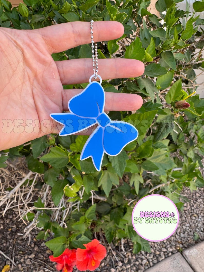 May include: A blue bow-shaped charm with a white outline, hanging from a silver ball chain. The charm is held in front of a green leafy background. The text "DESIGNS BY CRYSTAL" is visible.