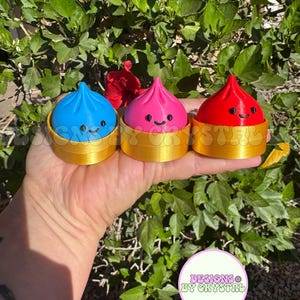 May include: Three colorful, smiling dumpling-shaped objects in blue, pink, and red, each sitting in a gold-colored basket. The objects have black eyes and a smiling mouth. The background is a lush green plant with a red flower. The image includes a logo that says "DESIGNS BY CRYSTAL".