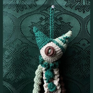 May include: A crocheted creature ornament featuring a pointed head with dark green and light green yarn, a single brown button eye, and a ruffled collar. The body is constructed from light green, dark green, and brown yarn, with a hanging loop.