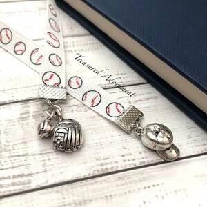 May include: White ribbon bookmark with a baseball theme. The ribbon has a repeating pattern of white baseballs with red stitching on a white background. The bookmark has a silver baseball glove charm, a silver baseball charm, and a silver baseball cap charm.