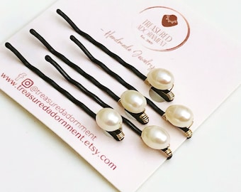 Puede incluir: Conjunto de seis pinzas para el pelo negras con detalles de perlas. Cada pinza tiene una pequeña perla blanca envuelta en alambre dorado. Las pinzas se muestran en una tarjeta blanca con el texto "Treasured Adornment" y una dirección de sitio web.