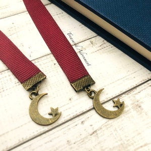 May include: Two red fabric bookmarks with a crescent moon and star charm. The bookmarks are on a white wooden surface. The text "Treasured Alignment" is visible on the surface.