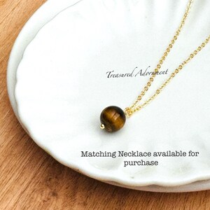 Pu&ograve; includere: Una collana con catena color oro con un pendente rotondo in pietra occhio di tigre marrone. La collana &egrave; esposta su un piatto bianco con bordo smerlato. Il testo sul piatto recita "Treasured Adornment" e "Matching Necklace available for purchase."