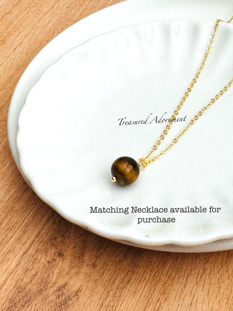 Pu&ograve; includere: Una collana con catena color oro con un pendente rotondo in pietra occhio di tigre marrone. La collana &egrave; esposta su un piatto bianco con bordo smerlato. Il testo sul piatto recita "Treasured Adornment" e "Matching Necklace available for purchase."