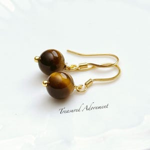 Pu&ograve; includere: Orecchini color oro con perline rotonde di occhio di tigre marroni. Gli orecchini hanno un design a gancio e il testo "Treasured Adornment" &egrave; visibile.