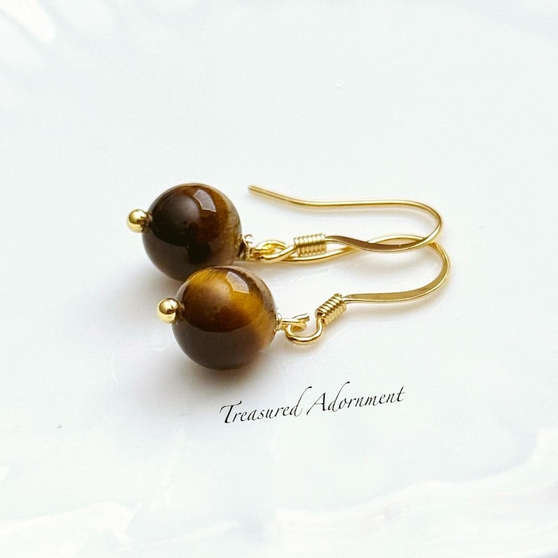 Pu&ograve; includere: Orecchini color oro con perline rotonde di occhio di tigre marroni. Gli orecchini hanno un design a gancio e il testo "Treasured Adornment" &egrave; visibile.